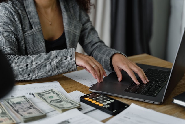 business-using-computer | Specialized Memphis Close up of a person using a laptop at a table with stacks of dollar bills and a phone calculator